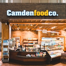 A small convenience store or food shop features a variety of snacks, drinks, and quick meals displayed on shelves, with a counter for service. The store has a wooden and brick design, with warm lighting hanging from the ceiling. Products like bottled water, chips, and packaged goods are visible.