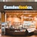 A small convenience store or food shop features a variety of snacks, drinks, and quick meals displayed on shelves, with a counter for service. The store has a wooden and brick design, with warm lighting hanging from the ceiling. Products like bottled water, chips, and packaged goods are visible.
