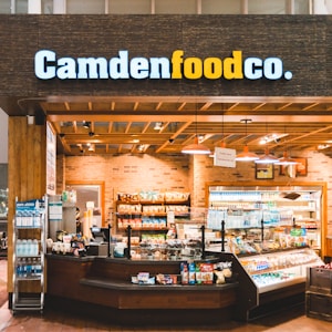 A small convenience store or food shop features a variety of snacks, drinks, and quick meals displayed on shelves, with a counter for service. The store has a wooden and brick design, with warm lighting hanging from the ceiling. Products like bottled water, chips, and packaged goods are visible.