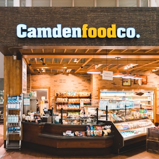 A small convenience store or food shop features a variety of snacks, drinks, and quick meals displayed on shelves, with a counter for service. The store has a wooden and brick design, with warm lighting hanging from the ceiling. Products like bottled water, chips, and packaged goods are visible.