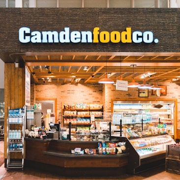 A small convenience store or food shop features a variety of snacks, drinks, and quick meals displayed on shelves, with a counter for service. The store has a wooden and brick design, with warm lighting hanging from the ceiling. Products like bottled water, chips, and packaged goods are visible.