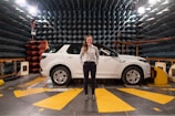 A woman stands confidently in front of a white SUV in an anechoic chamber with a patterned, soundproof design on the walls. The environment suggests a research or testing facility with technical equipment visible around the vehicle.