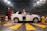 A woman stands confidently in front of a white SUV in an anechoic chamber with a patterned, soundproof design on the walls. The environment suggests a research or testing facility with technical equipment visible around the vehicle.