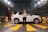 A woman stands confidently in front of a white SUV in an anechoic chamber with a patterned, soundproof design on the walls. The environment suggests a research or testing facility with technical equipment visible around the vehicle.
