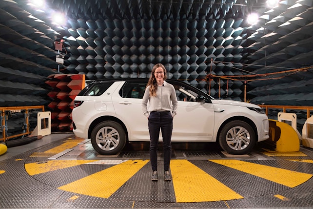 A woman stands confidently in front of a white SUV in an anechoic chamber with a patterned, soundproof design on the walls. The environment suggests a research or testing facility with technical equipment visible around the vehicle.