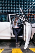 A person is sitting in a white vehicle with the door open, working on a laptop. The background has anechoic chamber panels, indicating a testing or experimental environment.