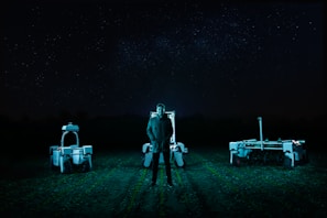 A person stands in a field at night, flanked by two robotic machines. The background is a dark night sky filled with stars. The scene is illuminated by cool-toned, artificial lighting, casting an eerie glow over the landscape.