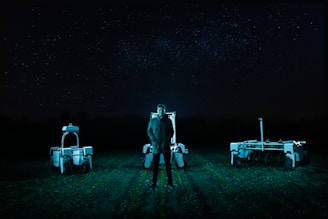 A person stands in a field at night, flanked by two robotic machines. The background is a dark night sky filled with stars. The scene is illuminated by cool-toned, artificial lighting, casting an eerie glow over the landscape.
