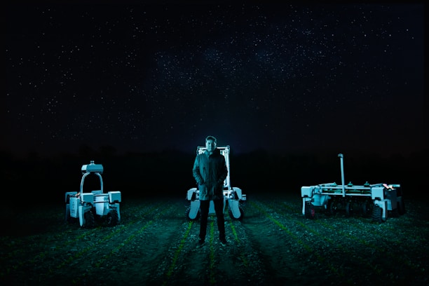 A person stands in a field at night, flanked by two robotic machines. The background is a dark night sky filled with stars. The scene is illuminated by cool-toned, artificial lighting, casting an eerie glow over the landscape.