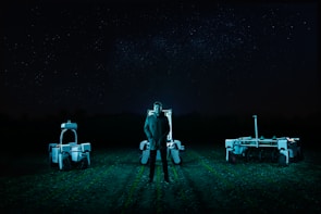 A person stands in a field at night, flanked by two robotic machines. The background is a dark night sky filled with stars. The scene is illuminated by cool-toned, artificial lighting, casting an eerie glow over the landscape.