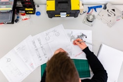 A person is working at a desk with technical drawings and a caliper. The desk contains various tools and equipment, including a toolbox labeled 'Stanley', a notebook, a cutting mat, and assorted mechanical parts and components.