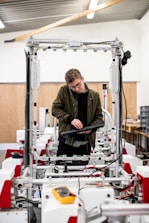 A man is standing in a workshop or industrial setting, interacting with a large piece of machinery or equipment. He is holding a keyboard and appears to be concentrating on the work at hand. The environment looks clean and organized, with various wires and components visible around him.