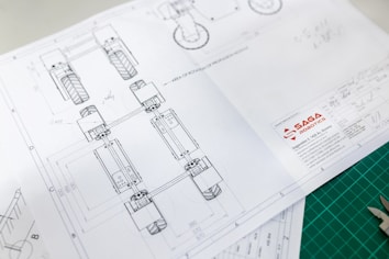 Technical drawing on white paper with detailed design layout, including geometric shapes and numbers, showing part of a mechanical structure. The design has annotations and measurements. The logo of 'SAGA Robotics' appears prominently on the lower right corner of the paper.