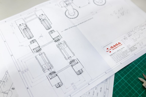 Technical drawing on white paper with detailed design layout, including geometric shapes and numbers, showing part of a mechanical structure. The design has annotations and measurements. The logo of 'SAGA Robotics' appears prominently on the lower right corner of the paper.