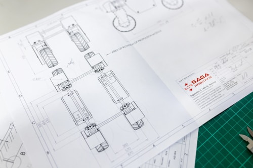 Technical drawing on white paper with detailed design layout, including geometric shapes and numbers, showing part of a mechanical structure. The design has annotations and measurements. The logo of 'SAGA Robotics' appears prominently on the lower right corner of the paper.