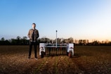 A person stands in a field next to an agricultural robot. The field has rows of young plants, and the horizon shows a soft gradient of blue and warm tones from an early sunset or sunrise. Trees line the edge of the field under the vibrant sky.
