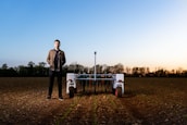 A person stands in a field next to an agricultural robot. The field has rows of young plants, and the horizon shows a soft gradient of blue and warm tones from an early sunset or sunrise. Trees line the edge of the field under the vibrant sky.