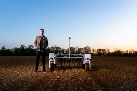 A person stands in a field next to an agricultural robot. The field has rows of young plants, and the horizon shows a soft gradient of blue and warm tones from an early sunset or sunrise. Trees line the edge of the field under the vibrant sky.