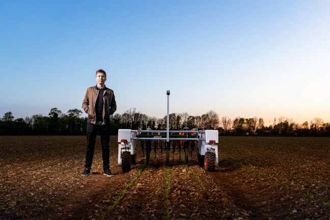 A person stands in a field next to an agricultural robot. The field has rows of young plants, and the horizon shows a soft gradient of blue and warm tones from an early sunset or sunrise. Trees line the edge of the field under the vibrant sky.
