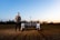 A person stands in a field next to an agricultural robot. The field has rows of young plants, and the horizon shows a soft gradient of blue and warm tones from an early sunset or sunrise. Trees line the edge of the field under the vibrant sky.