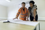 Two people stand at a table in a bright office. One wears an orange high-visibility jacket and gestures towards a large blueprint spread out before them, indicating a discussion or collaboration. The other person leans over the table, paying attention to the details on the paper.