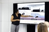 A person stands next to a large screen displaying diagrams and illustrations related to weirs and water management. They are gesturing towards the images, possibly explaining or presenting the information. The room appears to be a professional setting, with white walls and additional individuals seated and facing the screen.
