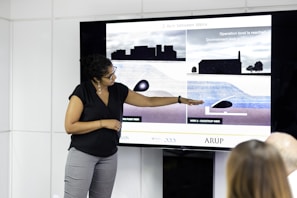 A person stands next to a large screen displaying diagrams and illustrations related to weirs and water management. They are gesturing towards the images, possibly explaining or presenting the information. The room appears to be a professional setting, with white walls and additional individuals seated and facing the screen.