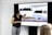 A person stands next to a large screen displaying diagrams and illustrations related to weirs and water management. They are gesturing towards the images, possibly explaining or presenting the information. The room appears to be a professional setting, with white walls and additional individuals seated and facing the screen.