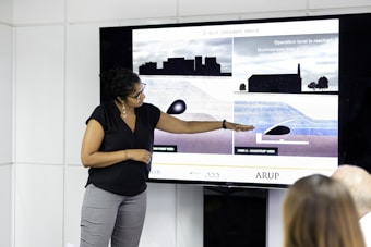 A person stands next to a large screen displaying diagrams and illustrations related to weirs and water management. They are gesturing towards the images, possibly explaining or presenting the information. The room appears to be a professional setting, with white walls and additional individuals seated and facing the screen.