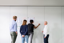 Four people stand in an office setting in front of a whiteboard or wall. Two individuals, one wearing a shirt and the other in a jacket, face the board where diagrams and notes are pinned. A person is actively writing or drawing with a marker while others observe.
