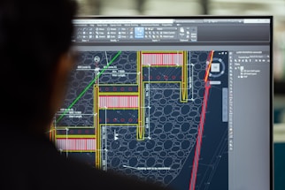 An individual is in front of a computer monitor displaying a detailed architectural or engineering design, possibly a floor plan or structural layout with measurements and annotations. The design is composed of various lines in colors like yellow, red, and green against a dark background, and the interface resembles a CAD software.
