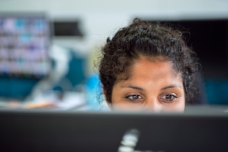 A person is looking over the top of a monitor, focusing intently. The background is blurred, showing a workplace setting with multiple monitors and documents.