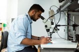 A person dressed in a light blue shirt is sitting on a black chair and looking through a microscope in a laboratory setting. The person appears focused, with one hand on the microscope and the other hand holding an object. Various laboratory equipment is visible around the individual in the well-lit room.