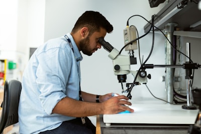 A person dressed in a light blue shirt is sitting on a black chair and looking through a microscope in a laboratory setting. The person appears focused, with one hand on the microscope and the other hand holding an object. Various laboratory equipment is visible around the individual in the well-lit room.