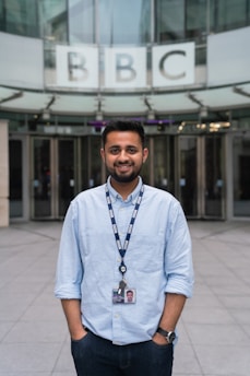 A person wearing a light blue shirt and a lanyard stands in front of a modern glass building entrance. The lanyard displays an ID card, and the letters 'BBC' are prominently displayed above the entrance.