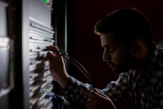 A person is concentrated on working with cables connected to a server rack in a dimly lit environment. The focus is on their hands and the server equipment, suggesting a technical task.