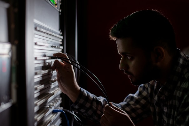 A person is concentrated on working with cables connected to a server rack in a dimly lit environment. The focus is on their hands and the server equipment, suggesting a technical task.