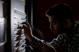 A person is concentrated on working with cables connected to a server rack in a dimly lit environment. The focus is on their hands and the server equipment, suggesting a technical task.