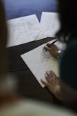 A person is working on architectural blueprints on a table, using a pen to make adjustments. Several sheets with technical drawings are spread out, indicating a focus on planning or design work.