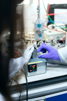 Two individuals wearing protective gloves are manipulating a small piece of equipment near a digital controller on a lab bench. The background is filled with wires and scientific apparatus suggesting a laboratory setting.