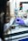 Two individuals wearing protective gloves are manipulating a small piece of equipment near a digital controller on a lab bench. The background is filled with wires and scientific apparatus suggesting a laboratory setting.