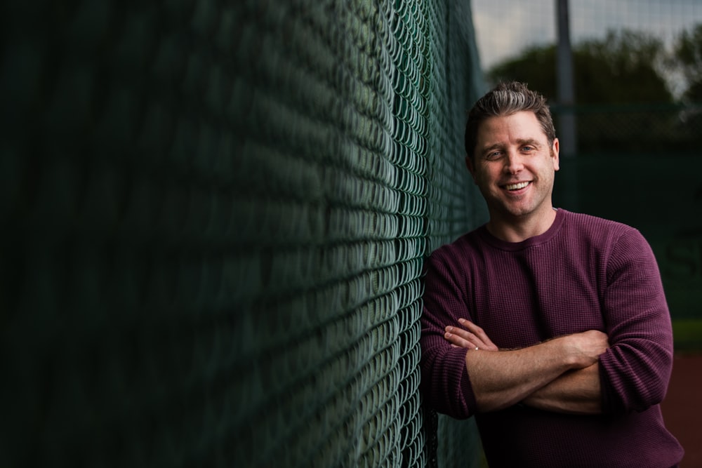 man in maroon crew neck t-shirt leaning on green metal fence during daytime