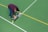 A man is bending over on a sports court, operating a line marking machine to draw straight lines. The court surface is green with white and yellow lines, indicating it may be used for multiple sports.