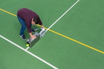 A man is bending over on a sports court, operating a line marking machine to draw straight lines. The court surface is green with white and yellow lines, indicating it may be used for multiple sports.