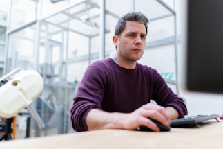 A man is focused on his work, seated at a desk in an industrial or laboratory setting. He is wearing a purple sweater and appears to be engaged with a task on the computer. The background includes lab equipment and glass structures, suggesting a scientific or tech environment.