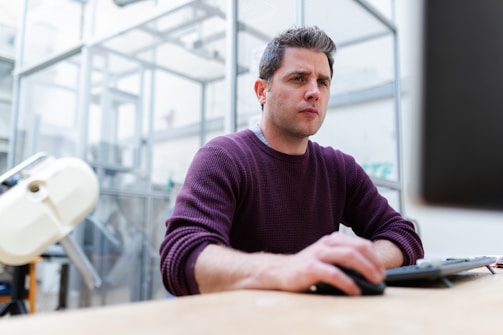 A man is focused on his work, seated at a desk in an industrial or laboratory setting. He is wearing a purple sweater and appears to be engaged with a task on the computer. The background includes lab equipment and glass structures, suggesting a scientific or tech environment.