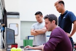 Three people are gathered around a desk, with one person seated and using a computer while the other two stand nearby and observe. The atmosphere appears informal and collaborative.