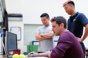 Three people are gathered around a desk, with one person seated and using a computer while the other two stand nearby and observe. The atmosphere appears informal and collaborative.