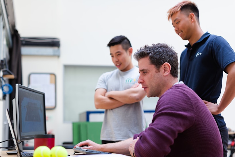 Three people are gathered around a desk, with one person seated and using a computer while the other two stand nearby and observe. The atmosphere appears informal and collaborative.