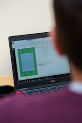 A person is looking at a laptop screen that displays a software interface. The screen shows a graphical layout or design, possibly related to a floor plan or technical drawing. There is also a black coffee mug next to the computer. The person appears to be seated in front of the laptop, focusing on the screen.